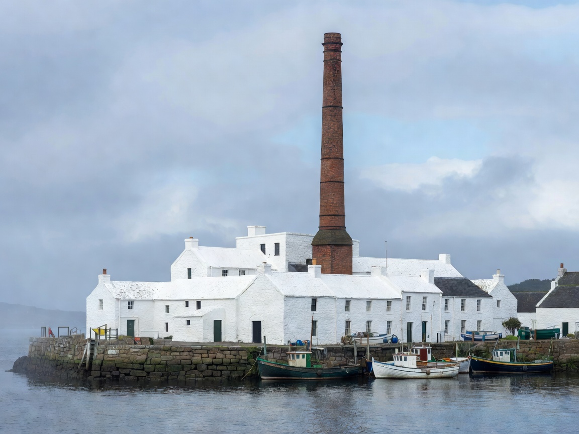 Whitewashed distillery with a tall brick chimney beside a small harbour in Campbeltown
